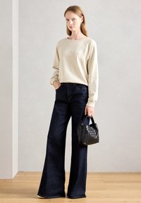 Felpa beige con logo ricamato, jeans a gamba larga blu scuro e una piccola borsa intrecciata nera con manico. Pavimento in legno e sfondo neutro.