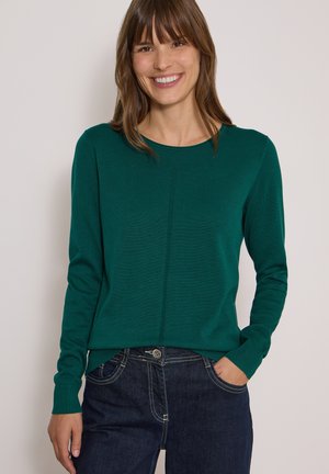 Femme souriante aux cheveux bruns portant un pull vert à manches longues et un jean bleu foncé, une main dans la poche.