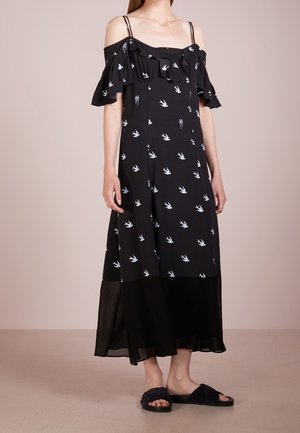 Robe midi noire épaules dénudées avec imprimé d'oiseaux blancs, détail à volants, fines bretelles, et ourlet transparent, portée avec des sandales noires à enfiler.