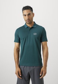Vyras su trumpais juodais plaukais, dėvintis tyliai mėlyną EA7 Emporio Armani polo marškinėlį ir pilkas šortus, stovintis prieš paprastą baltą foną.