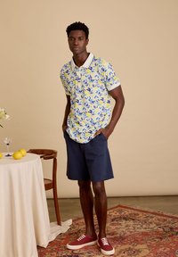 Korte mouwen polo in geel met blauwe citroen- en bladprint, gecombineerd met marineblauwe shorts en rode sneakers. Plaatje tegen een neutrale achtergrond.
