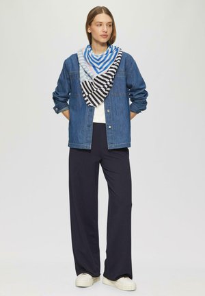 Vrouw draagt een blauw spijkerjasje, blauw-zwart gestreepte sjaal, wit shirt, zwarte wijde broek en witte sneakers, staat met haar handen achter haar rug.