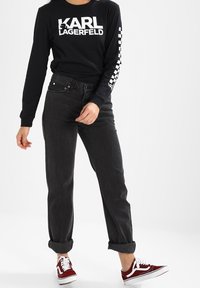 Camisa negra de manga larga con texto blanco, combinada con jeans oscuros de cintura alta y zapatillas burdeos con cordones y detalles blancos.