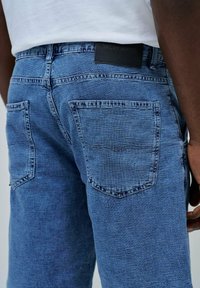 Pantalones vaqueros de denim en un lavado azul medio, con dos bolsillos traseros, tejido texturado y una etiqueta de marca en la cintura. Diseño clásico.