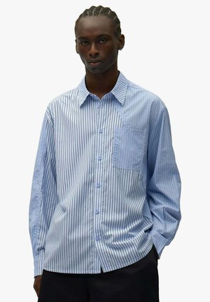 Giovane con i capelli intrecciati che indossa una camicia a righe verticali blu e bianche dal taglio ampio, con taschino sul petto.