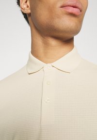 Gros plan d'une personne portant un polo texturé couleur crème avec un col et trois boutons fermés.