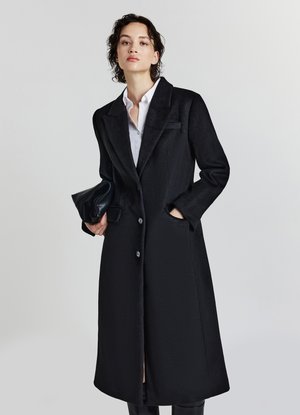 Cappotto nero in lana con design sartoriale, collo a revers, chiusura con due bottoni, tasche laterali e una texture liscia. Accompagnato da una pochette.