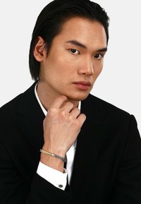 Bracciale argento e oro con design intrecciato, indossato su una mano appoggiata a un mento. Modello vestito con un completo nero e camicia bianca.