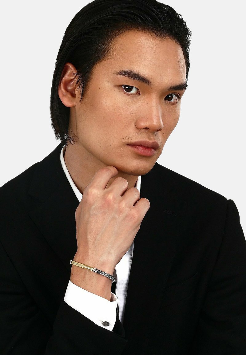 Bracciale argento e oro con design intrecciato, indossato su una mano appoggiata a un mento. Modello vestito con un completo nero e camicia bianca.