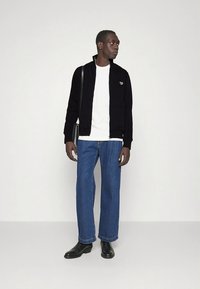 Veste noire zippée portée sur une chemise blanche, associée à un jean large bleu et des bottes noires. Le mannequin pose avec un sac à bandoulière.