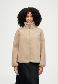 Tan fleece jacket with a high collar and zip closure. Features a textured surface, long sleeves, and contrasting seam accents.