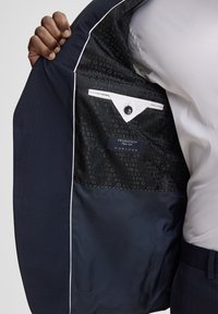 Das Innere eines Marineblazers mit glattem Futter, dezentem Markenmuster, weißem Taschenbesatz und Knopfverschluss.