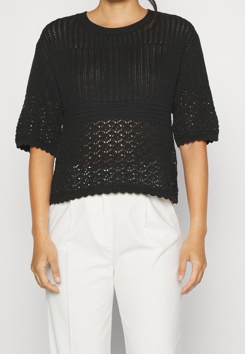 Svart stickad kortärmad topp med ett öppet vävmönster, ribbad halsringning och elastisk nederkant, bärs tillsammans med ljusa byxor.