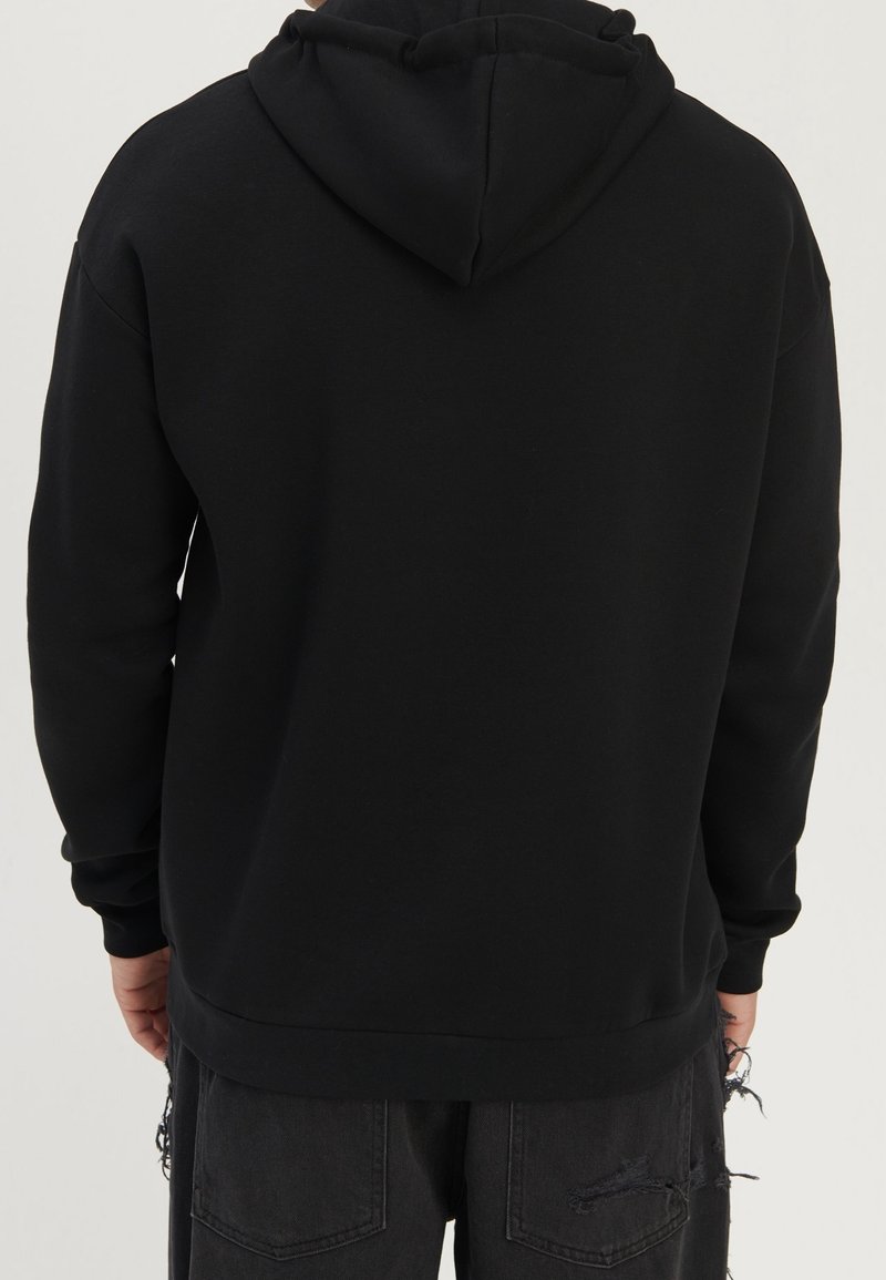 Personne portant un sweat à capuche noir uni et un jean noir déchiré, vue de dos sur un fond neutre.