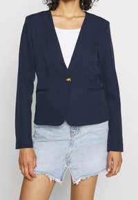 Blazer navy avec un décolleté en V profond, fermeture à un bouton doré, et deux poches avant. La coupe ajustée et la texture lisse s'associent parfaitement à une jupe en jean.