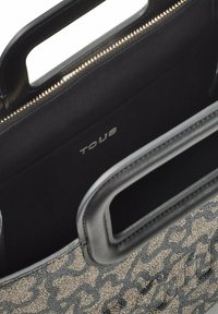 Bolso de mano negro y beige con un diseño estampado, manijas recortadas y un bolsillo interior con cremallera visible etiquetado "TOUS."