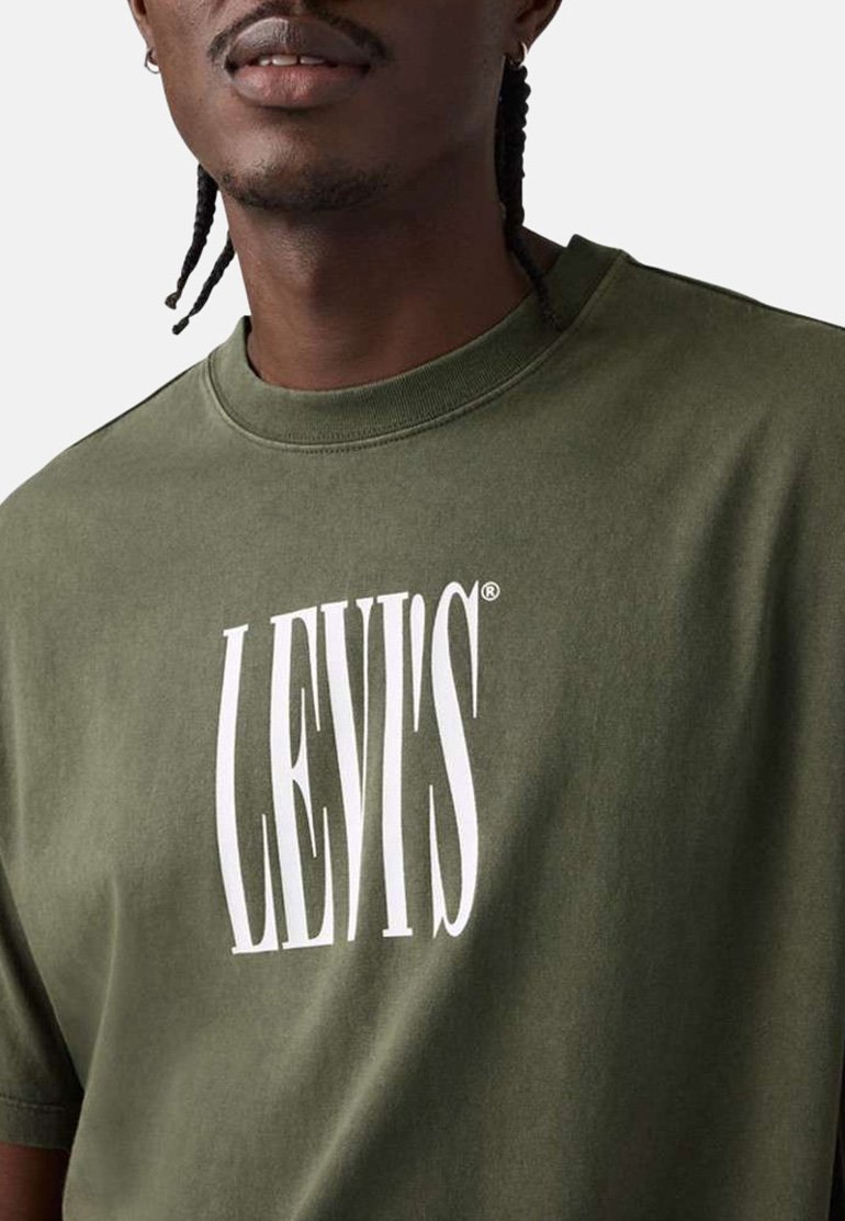 Hombre con camiseta verde oliva con el logo blanco distorsionado de "LEVI'S" en el frente, cabello trenzado y pequeño aro en la oreja izquierda.