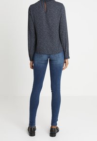 Blusa a maniche lunghe di colore blu navy con un motivo sottile, caratterizzata da un dettaglio a chiave sul retro, abbinata a jeans blu aderenti e scarpe nere.