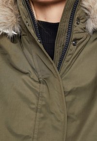 Parka verde oliva con cappuccio foderato in pelliccia, dotata di chiusura con zip, tessuto morbido e dettagli di cucitura a vista. Nessuna tasca visibile.