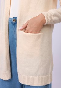 Cardigã longo bege claro com punhos e barra canelados, apresentando um bolso frontal e tecido suave e texturizado; combinado com jeans azuis.