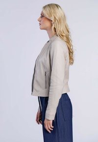 Ljust beige läderjacka med en slank, åtsittande design, sidoficka med dragkedja och ribbade muddar; matchad med marinblå byxor.