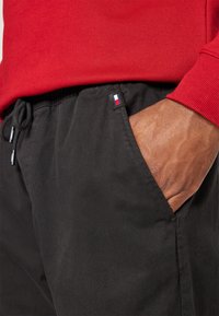 Svarta bomullsjoggers med elastisk midja och snöre, med en liten röd, vit och blå logotyp på fickan. Röd sweatshirt synlig.