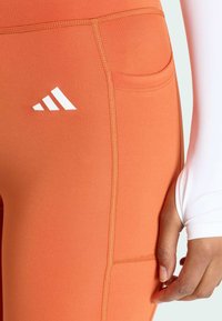 Oranžové športové legíny s bočnou kapsou. Vyrobené z elastického materiálu, s viditeľným šitím a bielym logom Adidas ako akcentom.