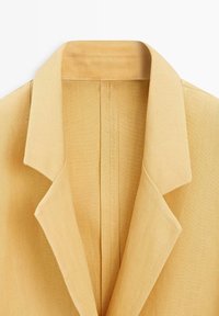 Gelber Leinenblazer mit Kragen, gekerbten Revers und sichtbaren Steppnähten auf weißem Hintergrund.