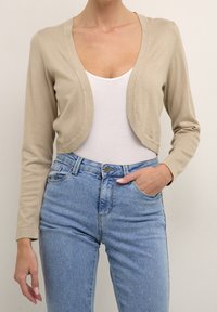 Beżowy krótki kardigan z lekkiego dzianiny, z głębokim dekoltem w serek i długimi rękawami, zestawiony z niebieskimi spodniami o wysokim stanie.