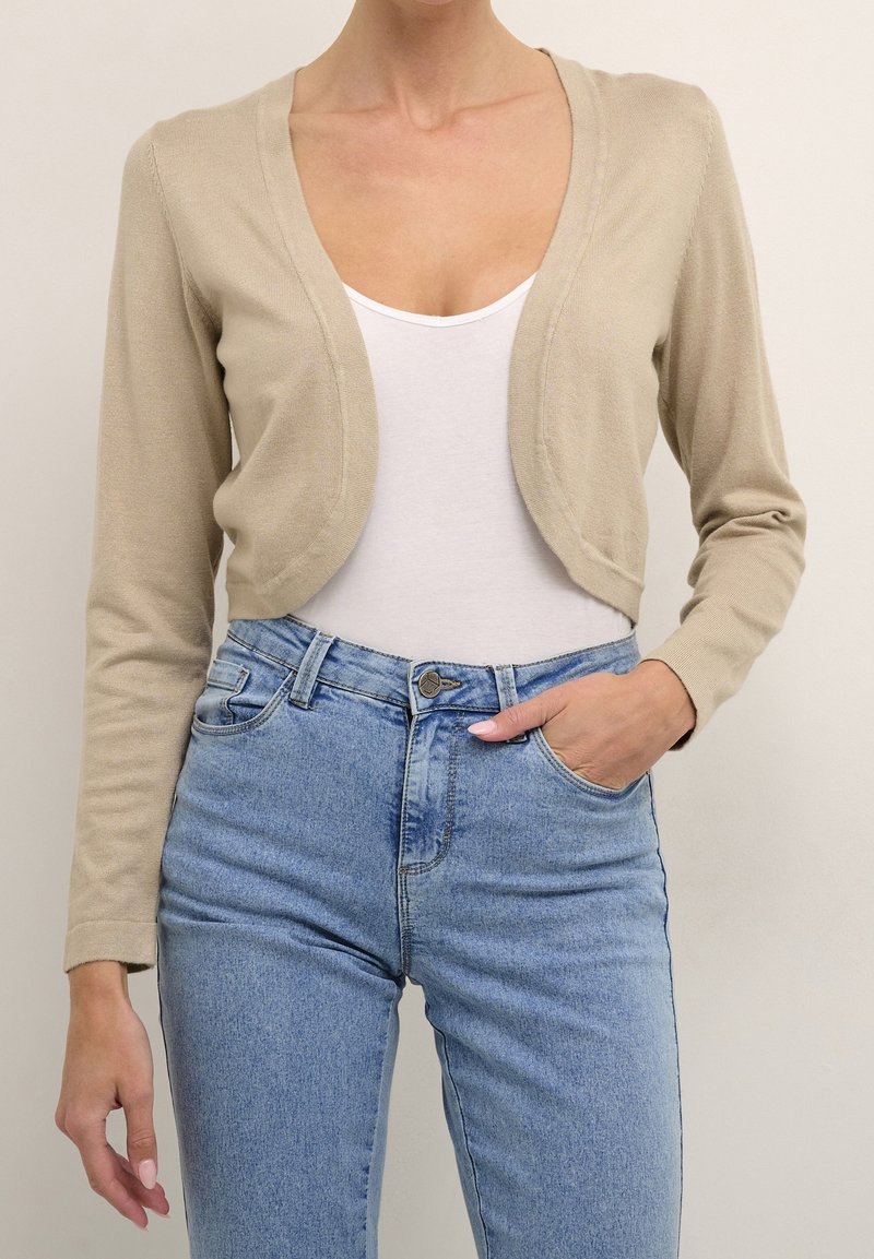 Beżowy krótki kardigan z lekkiego dzianiny, z głębokim dekoltem w serek i długimi rękawami, zestawiony z niebieskimi spodniami o wysokim stanie.