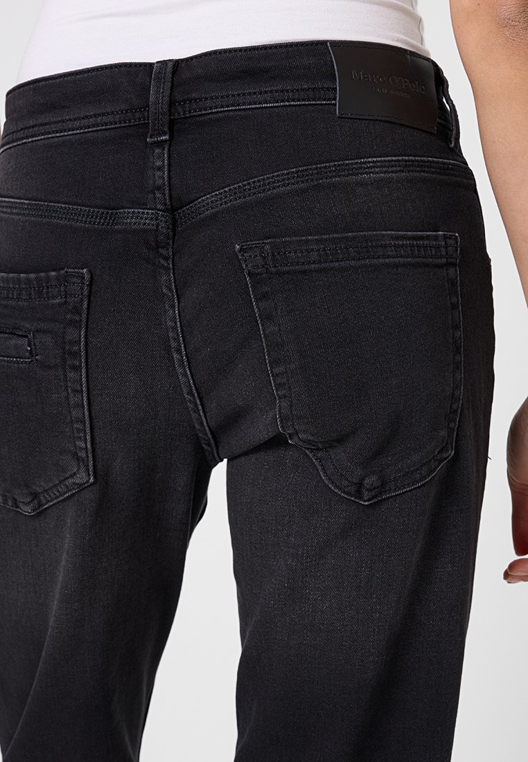 Widok z tyłu osoby w czarnych dżinsach z dużymi tylnymi kieszeniami i białej koszuli włożonej do spodni, na której widoczna jest skórzana metka marki.