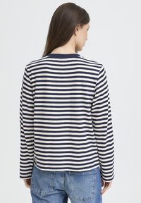 Pikkade pruunide juustega naine, kes kannab tumesinise ja valge triibulise pikkade varrukate ja siniste teksadega särki, vaadates selja tagant ühtlasel taustal.