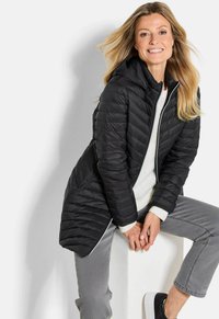 Vrouw met lang blond haar glimlacht, gekleed in een zwarte gewatteerde jas, witte trui, grijze jeans en zwarte instappers, zittend op een witte blok.