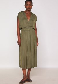 Vestito verde oliva, lungo fino al ginocchio, con scollatura a V, maniche corte e pieghe verticali. Indossato con scarpe ballerine marroni. Tessuto liscio.