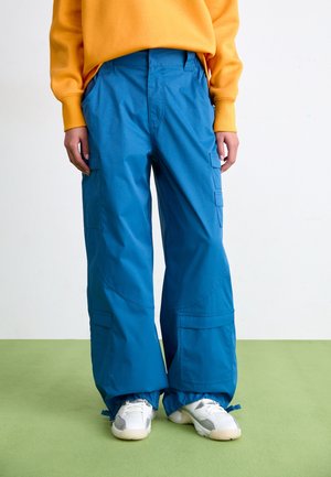 Personne portant un pantalon cargo bleu vif et des baskets blanches, debout sur un sol vert devant un fond blanc.