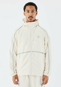 Chaqueta ligera de crema con ribetes grises, cierre completo y capucha ajustable. Cuenta con bolsillos frontales y puños elásticos. Textura de tela suave.