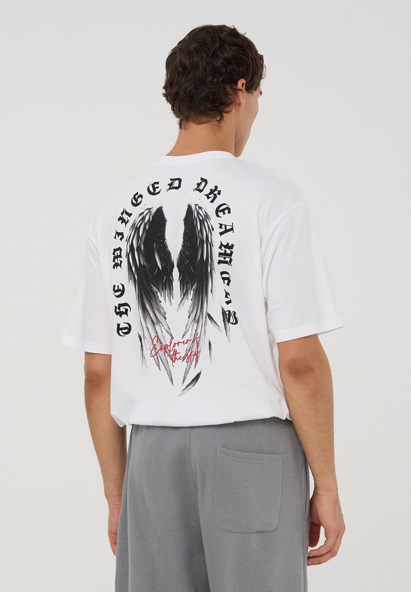 Maglietta di cotone bianco con una grande grafica di ali nere e grigie sul retro, con testo in tipografia in grassetto sopra e sotto il design.