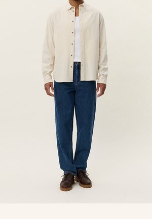 Mand iført beige skjorte med knapper over hvid tanktop, blå jeans og brune lædersko, stående mod en ensfarvet lys baggrund.
