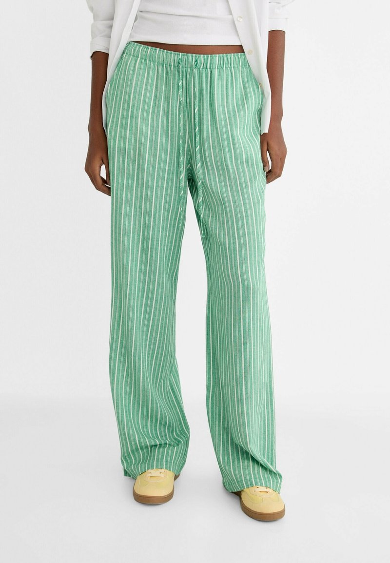 Personne portant un pantalon large rayé vertical vert et blanc avec taille élastique et chaussures jaunes, top blanc court et chemise.