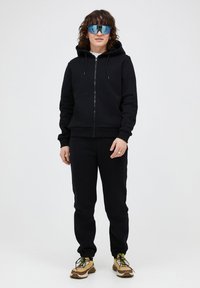 Zwarte zip-up hoodie met kangoeroe zak, gecombineerd met bijpassende zwarte trainingsbroek. Reflecterende blauwe zonnebril en stevige witte en gele sneakers.