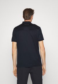 Polo noir avec un empiècement et un col texturés, à manches courtes, et un logo dans le dos, associé à un pantalon sombre.
