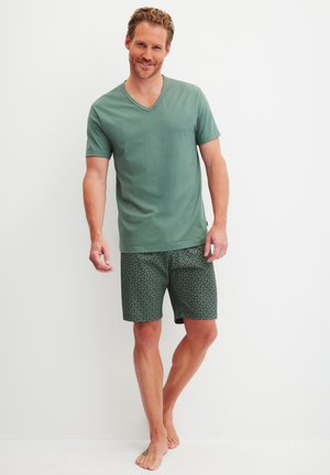 Groene korteglobige V-hals T-shirt gecombineerd met gemønsterde donkergroene shorts met cirkelvormige ontwerpen, model staat blootsvoets tegen een witte achtergrond.