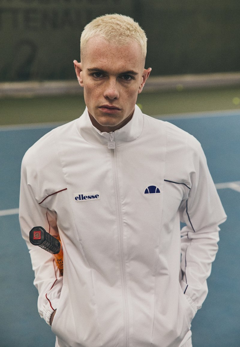 Weiße Sportjacke mit blauen Akzenten, Reißverschluss und Logo. Das Model hält einen Tennisschläger, im Hintergrund ist ein Platz mit einer grünen Wand zu sehen.