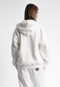 Sweatshirt à capuche blanche en tissu doux, doté d'une poche kangourou et d'un patch brodé rond en bas à gauche.