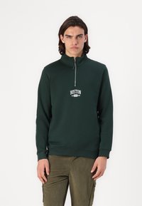 Mørkegrønn zip-up hettegenser med høy krage, med hvit tekst "BOSTON 19 USA 98", kombinert med olivengrønne kordfløyelsbukser.