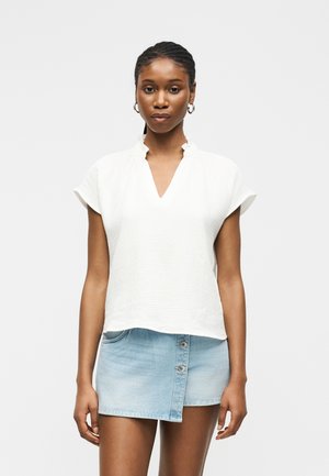 Femme aux cheveux tressés portant des boucles d’oreilles créoles dorées, un chemisier blanc à col à volants, et une jupe en denim asymétrique bleu clair.