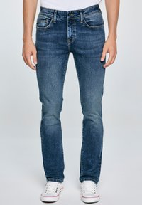 Jeans azules de mezclilla, con un corte ajustado, diseño estándar de cinco bolsillos, acabado desgastado y costuras en contraste, combinados con zapatillas blancas.