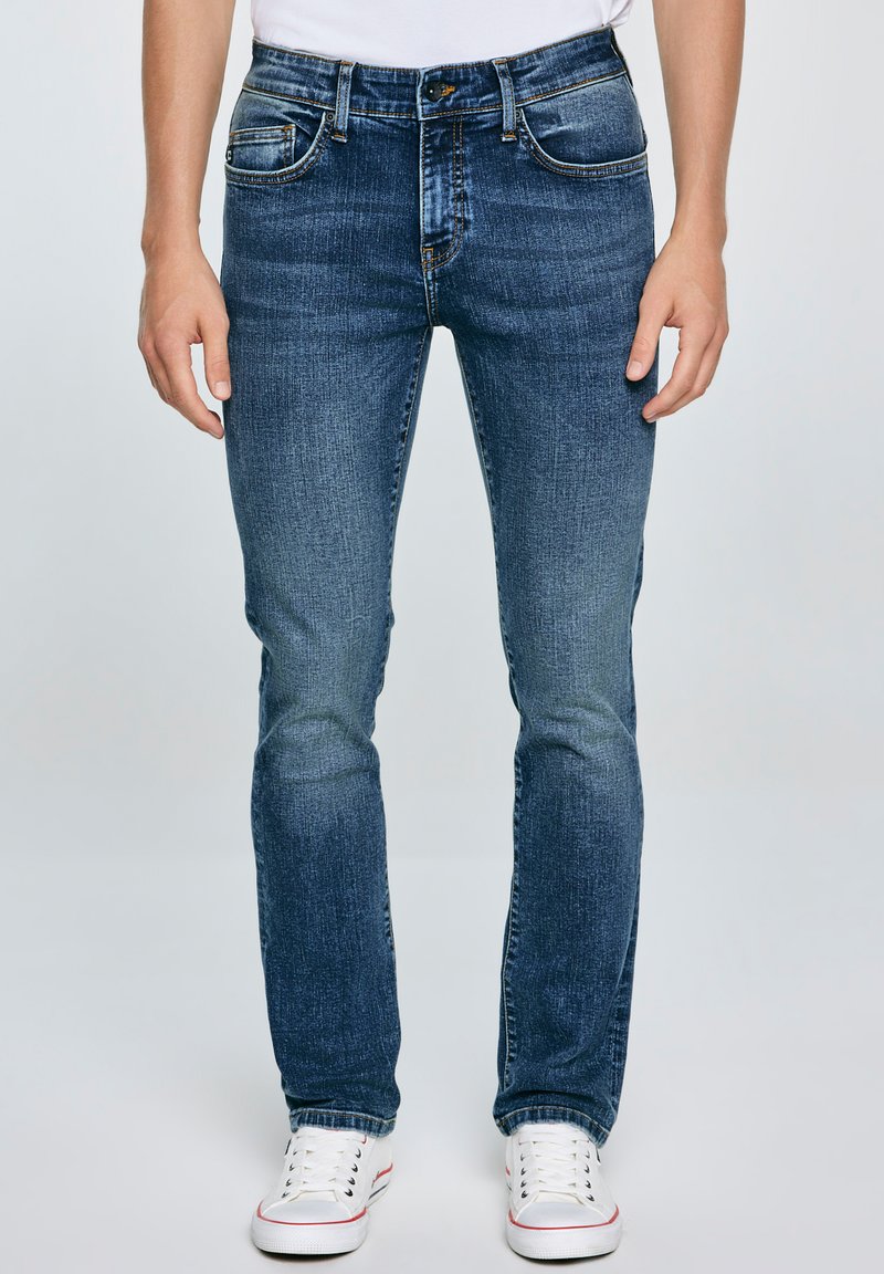 Jeans azules de mezclilla, con un corte ajustado, diseño estándar de cinco bolsillos, acabado desgastado y costuras en contraste, combinados con zapatillas blancas.