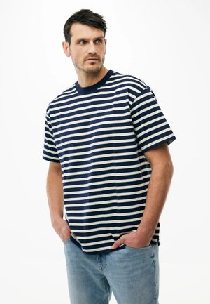 Mann mit kurzen dunklen Haaren, der ein marineblau-weiß gestreiftes T-Shirt und hellblaue Jeans trägt, steht mit den Händen in den Taschen vor weißem Hintergrund.