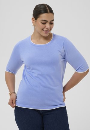 Femme portant un haut bleu clair à manches courtes avec bordure blanche et un jean foncé, souriante et regardant vers le bas, sur un fond clair uni.